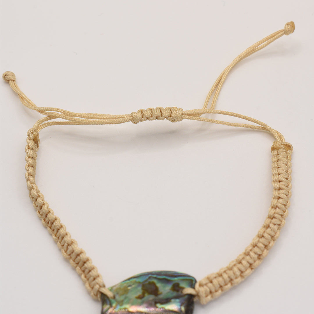 A close-up of the Salty Dreamers Bracelet | Paua Shell Rangi, featuring a handmade beige macrame band and a vibrant iridescent pāua shell centerpiece against a plain white background.