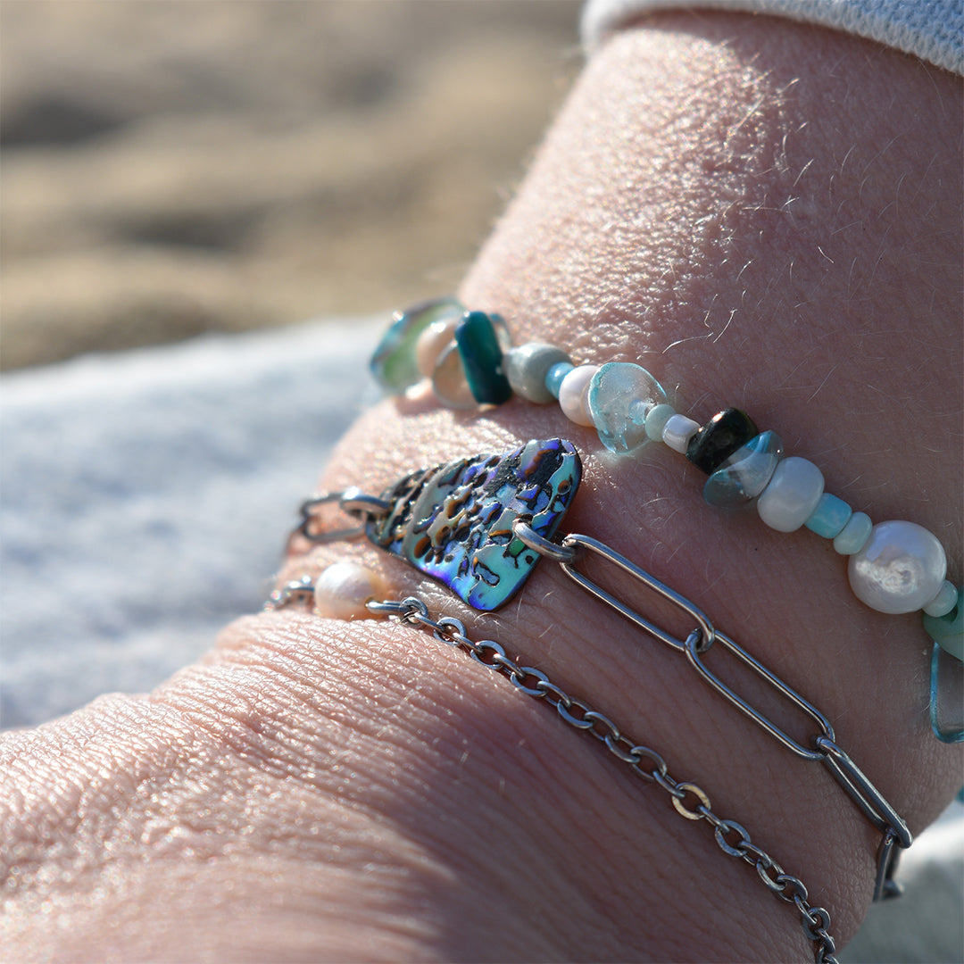 A close-up of a wrist wearing three bracelets, including the Salty Dreamers Bracelet | Ocean Bamboo Blue, set against a blurred outdoor background.
