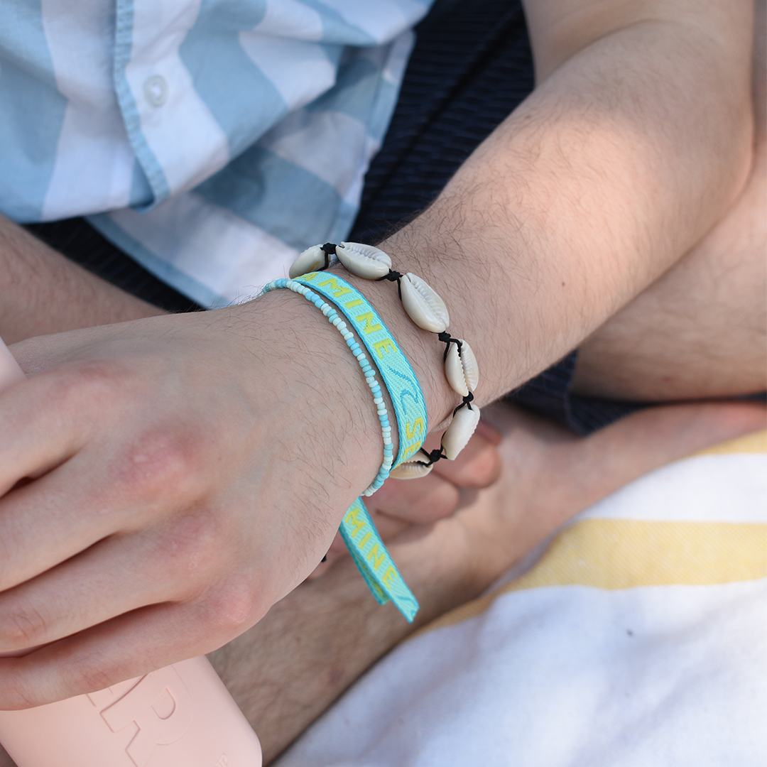 A person sits cross-legged in a blue and white plaid shirt, wearing the Salty Dreamers Bracelet | Cowrie Away Black. They hold a pale pink bottle, with a yellow and white striped towel nearby.