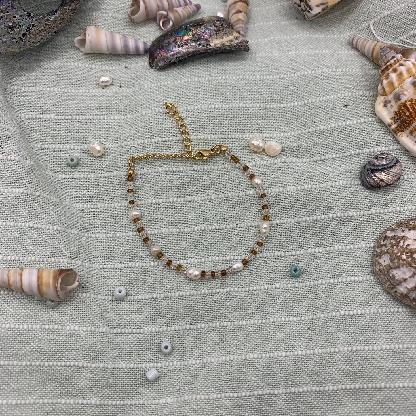 The Bracelet | caramel whispers by Salty Dreamers features gold, glass beads, and pearl-like accents. It’s showcased on a pale green textured cloth with assorted seashells and small beads.