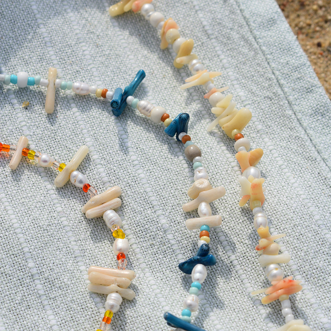 The Salty Dreamers Necklace | Ocean Bamboo Orange features handmade design with irregular shell pieces, blue coral, and colorful beads, displayed on light blue textured fabric with sand in the background.