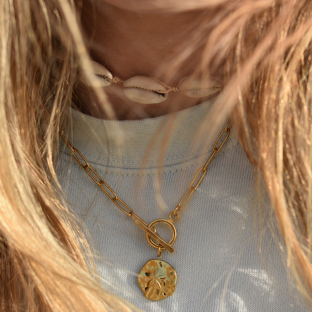 Close-up of a person with long, light brown hair in a white top, styled with beach jewelry: Salty Dreamers Necklace | Cowrie Choker and a gold chain necklace featuring a round, textured pendant.