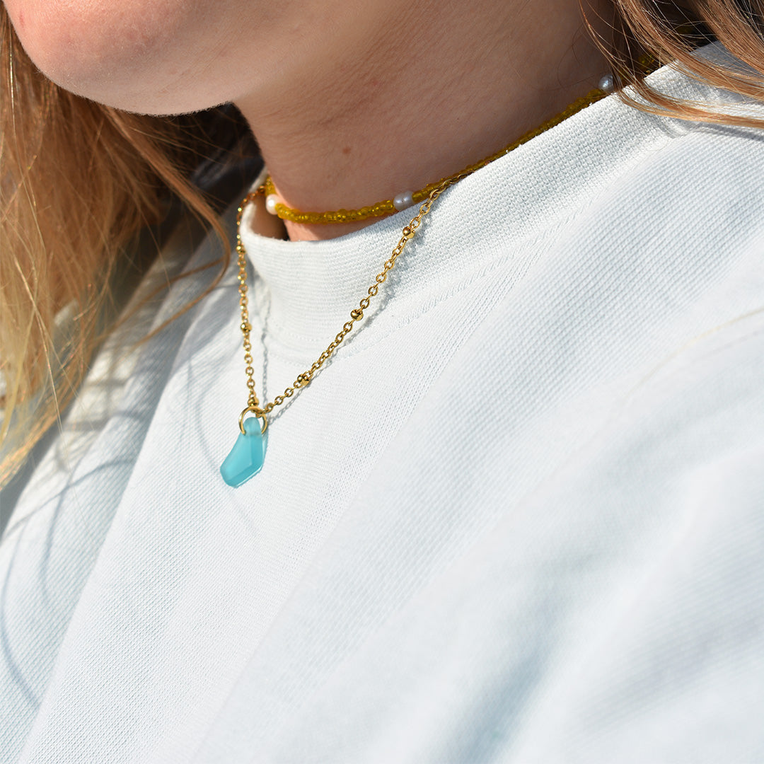 Close-up of a person in a white shirt wearing two gold necklaces—one featuring the Salty Dreamers Necklace | Starfish with a blue pendant—showcasing handcrafted colourful jewellery from Germany, with light brown hair partially visible.