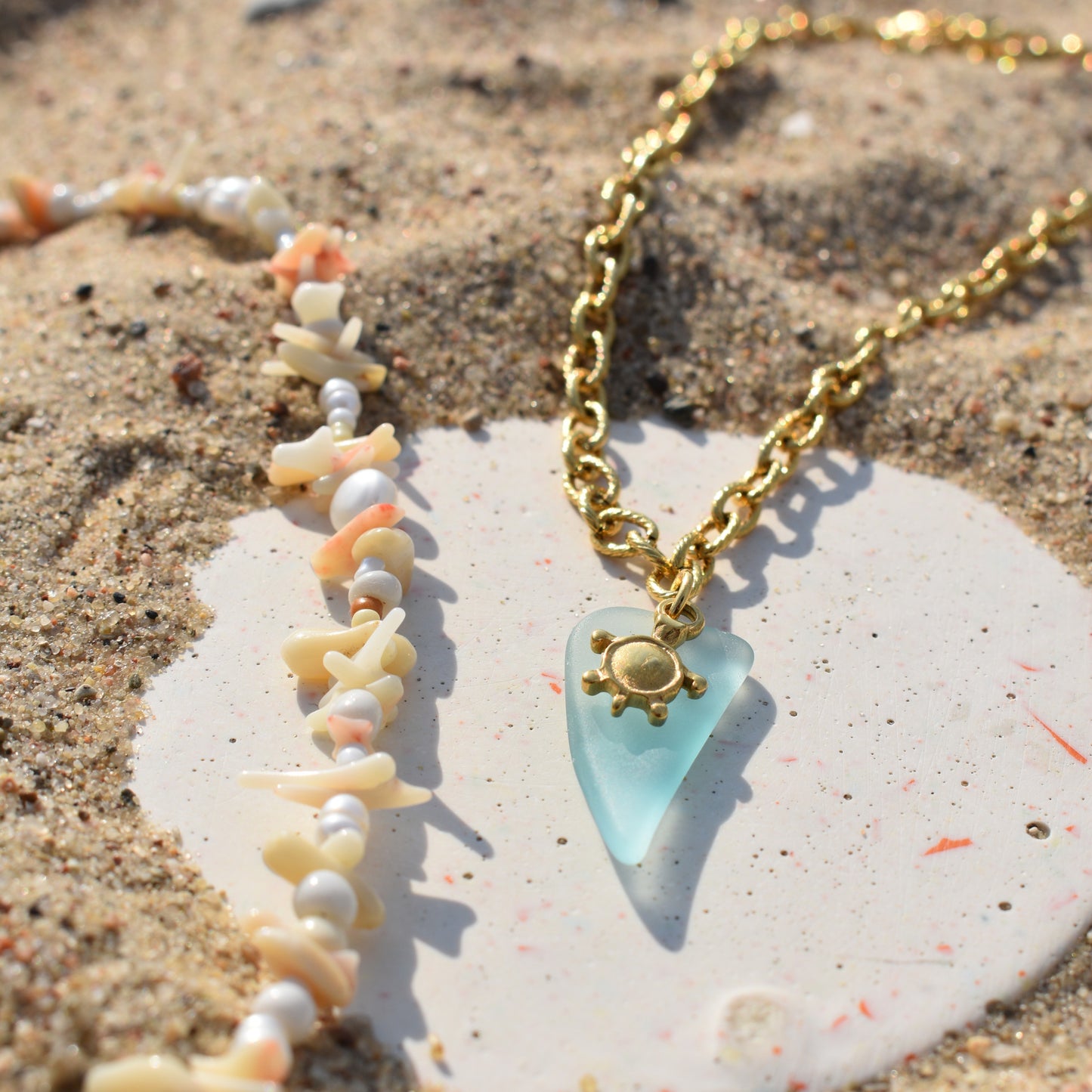 The Salty Dreamers Turtle Sea Glass Necklace rests on sandy ground beside a shell and bead bracelet, both displayed on a white, round stone.