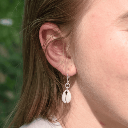 A close-up shows a person wearing Salty Dreamers' Earrings | Dangling Cowrie Silver—925 silver hoops with white cowrie shell pendants—standing out against a softly blurred green background.