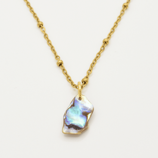 The Salty Dreamers Necklace | Paua Shell Ra features a gold chain with small round beads and an irregular iridescent pāua shell pendant—handmade jewelry shown on a plain white background.