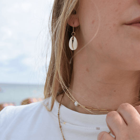 A woman wearing Salty Dreamers' Dangling Cowrie Gold earrings and a gold-plated 925-silver chain necklace with a white bead holds her necklace. The blurred background shows a beach and ocean scene.