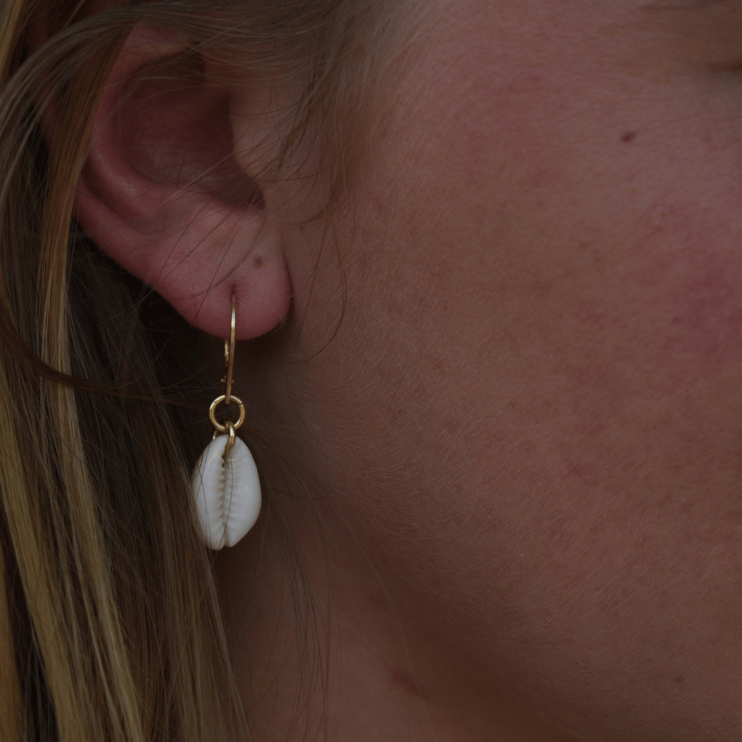 A close-up of a light-skinned, blonde-haired person wearing Salty Dreamers' Earrings | Dangling Cowrie Gold—stylish handmade gold-plated 925-silver hoops with white cowrie shell pendants.