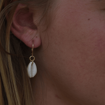 A close-up of a light-skinned, blonde-haired person wearing Salty Dreamers' Earrings | Dangling Cowrie Gold—stylish handmade gold-plated 925-silver hoops with white cowrie shell pendants.