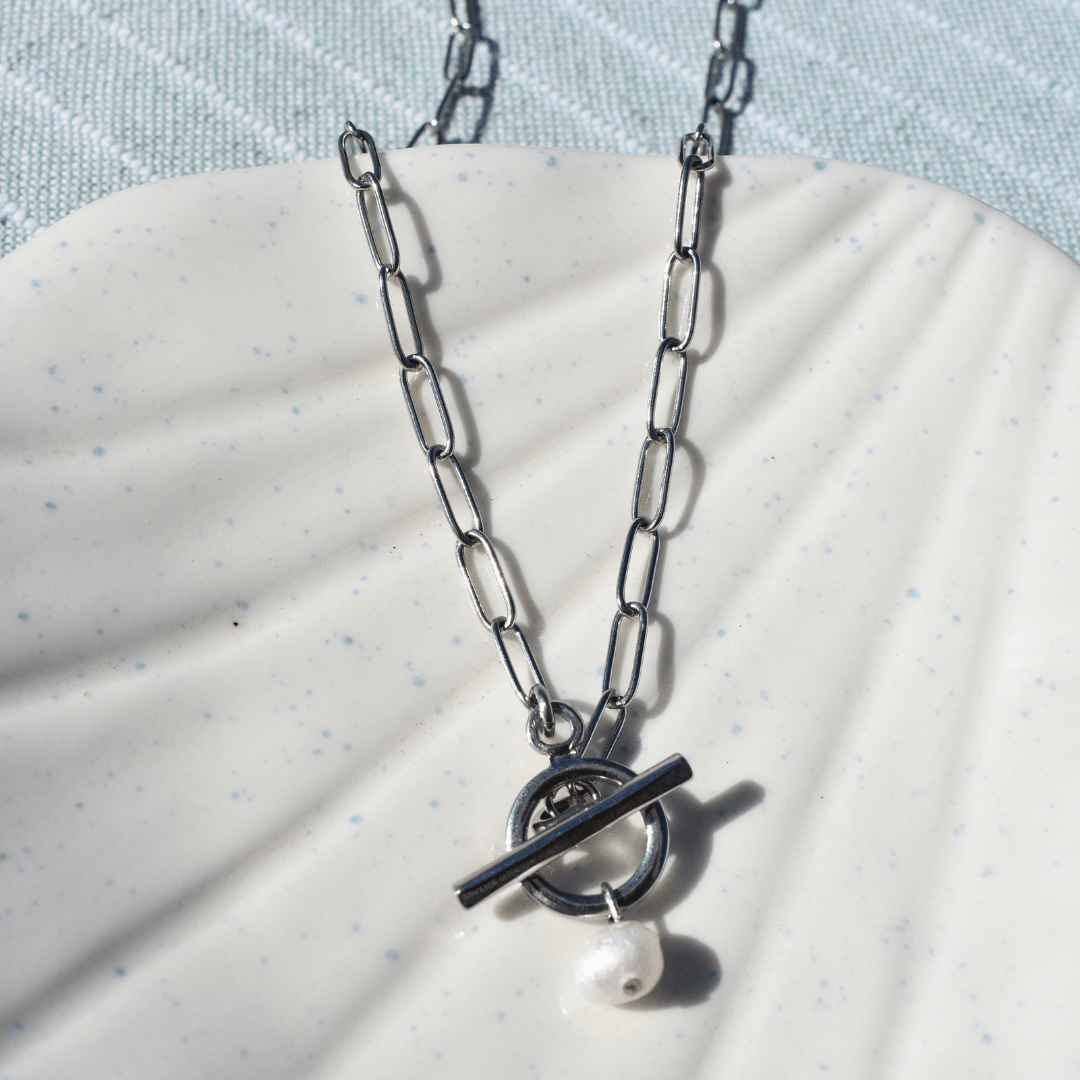 Salty Dreamers’ Necklace | The Cutie Pearl, featuring a handmade freshwater pearl on a silver chain with a toggle clasp, is displayed on a white shell-shaped dish against textured light blue fabric for a surfer-inspired vibe.