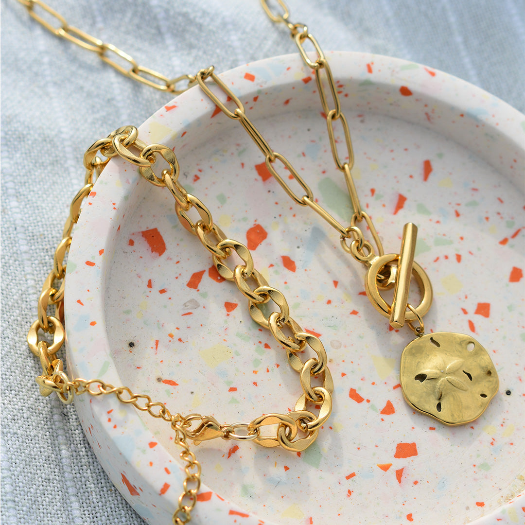 Zwei goldfarbene Salty Dreamers Ketten in einer runden Terrazzo-Schale, darunter das Stainless Waves Armband mit Sand-Dollar-Anhänger.