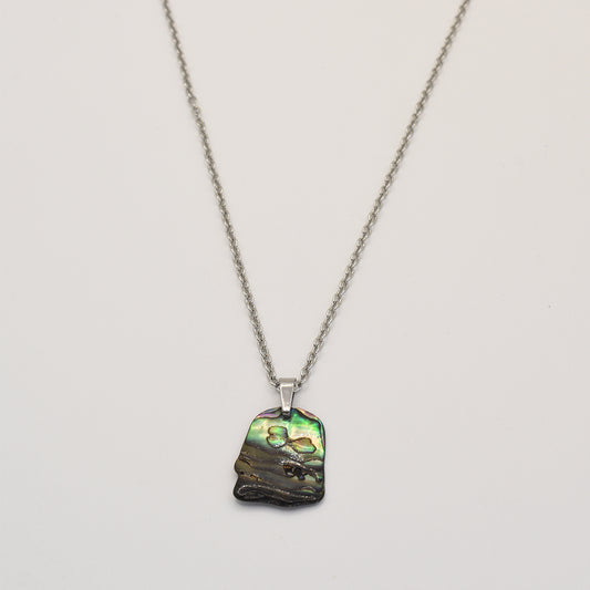 The Salty Dreamers Necklace | Paua Shell Kowhai features a handmade silver chain and an irregularly shaped, iridescent green and black pāua shell pendant, showcased on a plain white background.