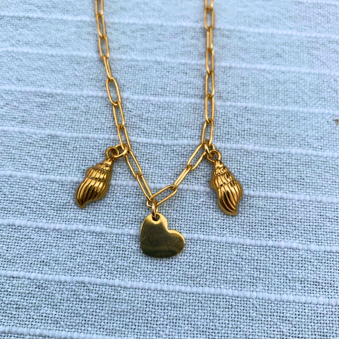 A gold chain necklace with two angel wing charms and a heart charm from Salty Dreamers' Workshop Meeresschmuck in Kiel, shown on light blue textured fabric.