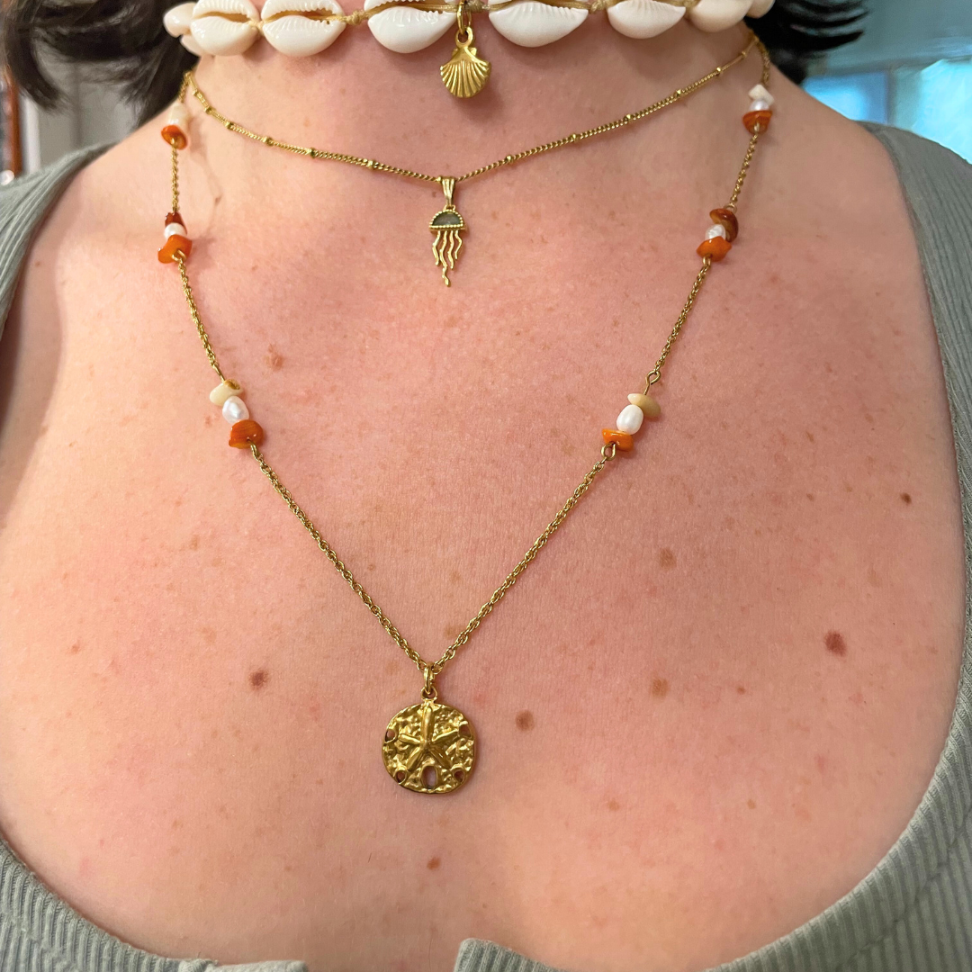 A person wears layered necklaces—a white shell choker, a gold seashell pendant, and a beaded gold chain—over a light green ribbed top. Perfect inspiration for Salty Dreamers' Workshop Ocean Jewellery in Kiel.