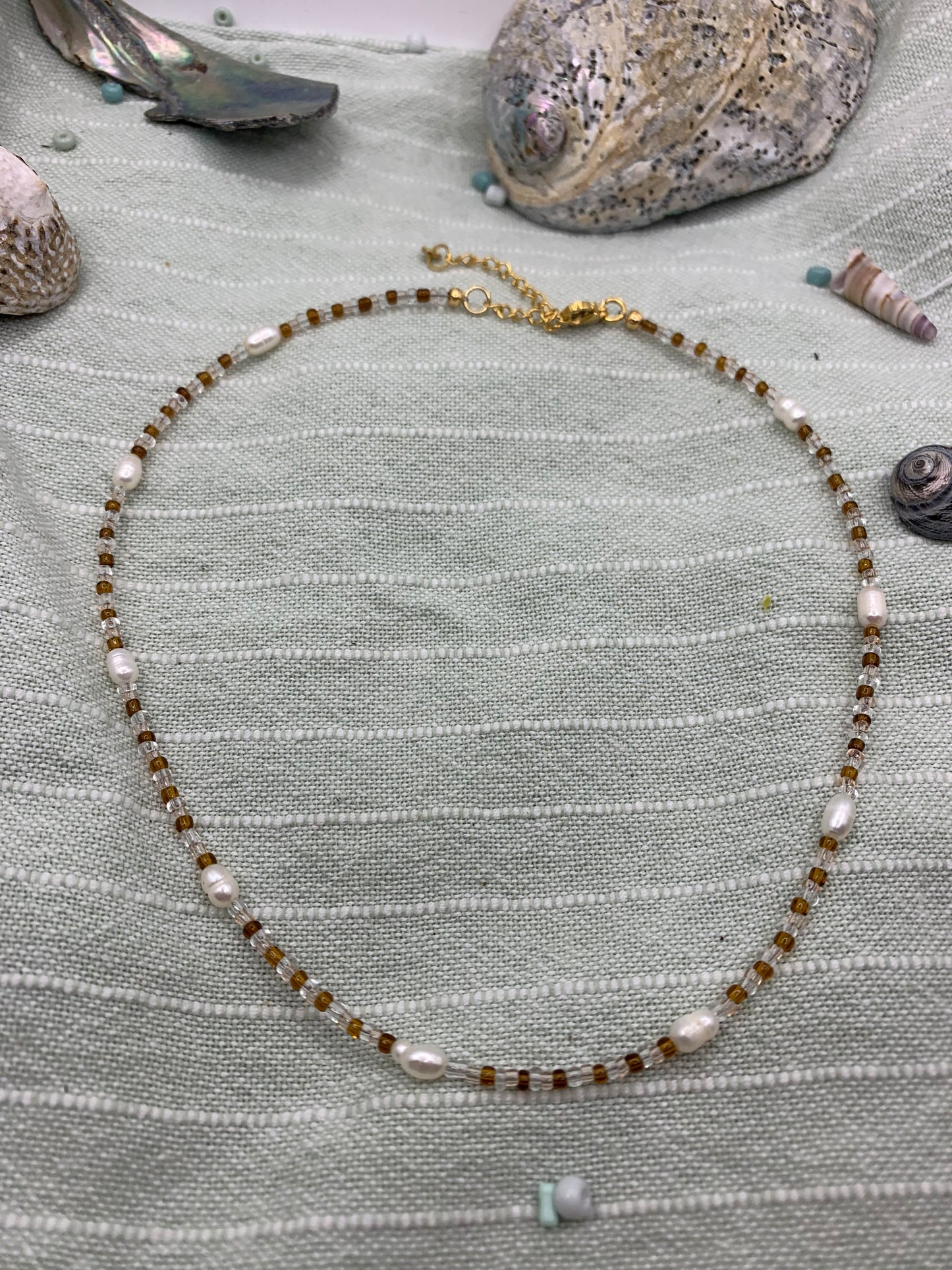 The Necklace | Caramel Whispers by Salty Dreamers features handmade brown and white freshwater pearls, elegantly displayed on light green striped fabric with seashells and stones—ideal for ocean lovers.