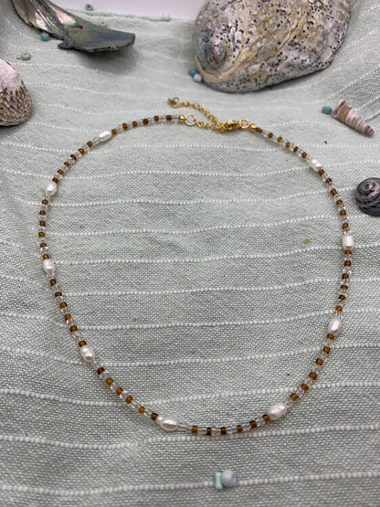 The Necklace | Caramel Whispers by Salty Dreamers features handmade brown and white freshwater pearls, elegantly displayed on light green striped fabric with seashells and stones—ideal for ocean lovers.