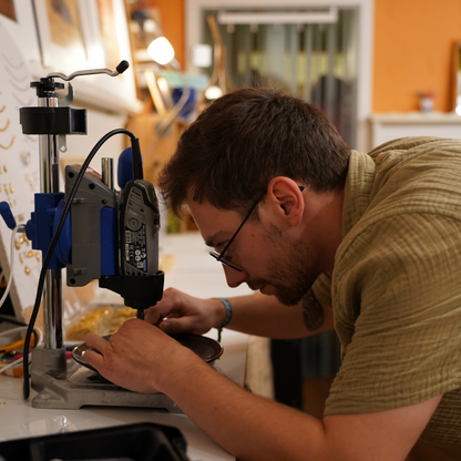 At the Workshop Ocean Jewellery in Kiel by Salty Dreamers, a man with glasses focuses intently as he uses a drill press on a small object at a well-lit workstation, creating unique jewelry in a cozy, creative studio.